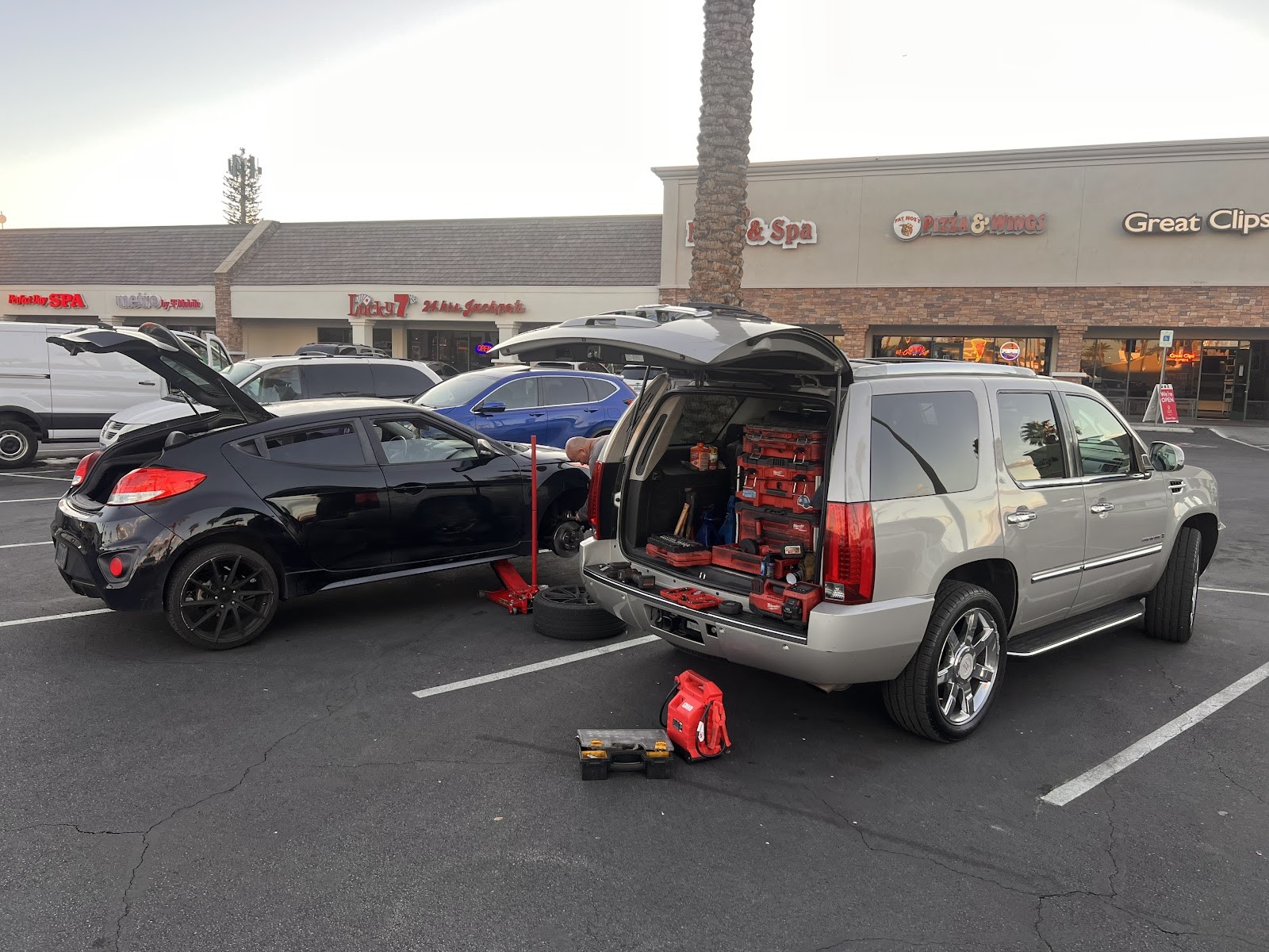 Mobile mechanic working roadside with a tool-loaded Escalade beside a car being serviced in a strip mall lot
